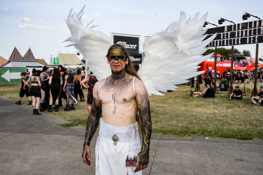 Dieses Bild zeigt einen Festivalbesucher mit kunstvollem Kostüm auf dem M'era Luna Festival.Veranstaltungsort: Das Foto wurde auf dem Flugplatz Drispenstedt in Hildesheim aufgenommen.Festivalcharakter: Das M'era Luna ist eines der größten Musikfestivals der schwarzen Szene in Deutschland.Kostüm: Der Besucher trägt auffällige weiße Flügel und ein detailliertes Make-up als Teil seines Festival-Outfits