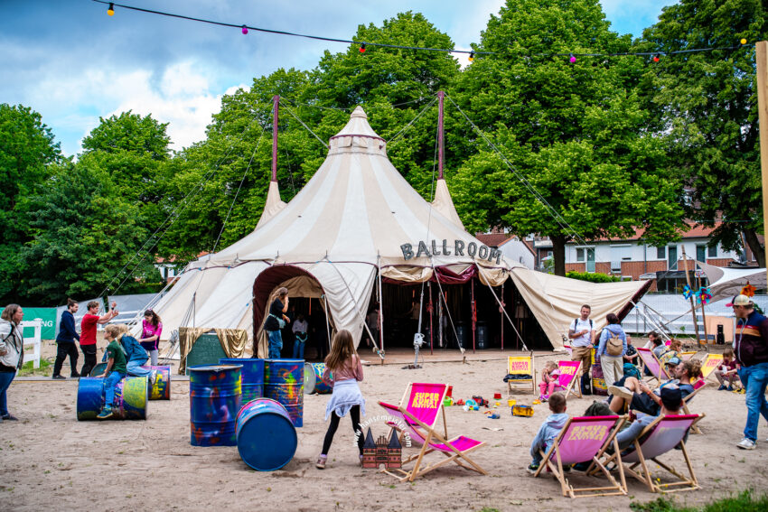 Hier ist die professionelle, atmosphärische Bildbeschreibung für deine Galerie bzw. deine Social-Media-Kanäle, inklusive der gekürzten Version und allen gewünschten Metadaten:

Festival-Impressionen: Der „Ballroom“ auf dem Super Kunst Festival 2026
Das Bild superkunstfestival-Freitag-12-06-2026 (68).jpg fängt die lebendige, einladende und urbane Atmosphäre des Festival-Freitags perfekt ein. Im Zentrum der Aufnahme steht ein großes, nostalgisches Rundzelt in Beige- und Weißtönen, das über dem Eingang stolz den Schriftzug „BALLROOM“ trägt. Das Zelt ist eingebettet in eine Kulisse aus dichten, grünen Bäumen unter einem leicht bewölkten Sommerhimmel. Eine quer durch das Bild gespannte Lichterkette mit bunten Glühbirnen deutet bereits auf die stimmungsvollen Abendstunden hin.

Der Vordergrund zeigt einen großzügigen, sandigen Publikumsbereich, der an eine urbane Strandbar erinnert. Hier verteilt stehen bunt mit Graffiti besprühte Ölfässer, die als Stehtische oder Dekoration dienen. Mehrere pinkfarbene Liegestühle mit dem unverkennbaren „Super Kunst Festival“-Branding laden zum Verweilen ein. Die Szene ist voller Leben: Besucher jeden Alters – darunter spielende Kinder auf den Fässern, entspannte Gruppen in den Liegestühlen und flanierende Festivalgäste – beleben den Platz vor dem Zelt und spiegeln den inklusiven, familiären und weltoffenen Charakter des Events wider.