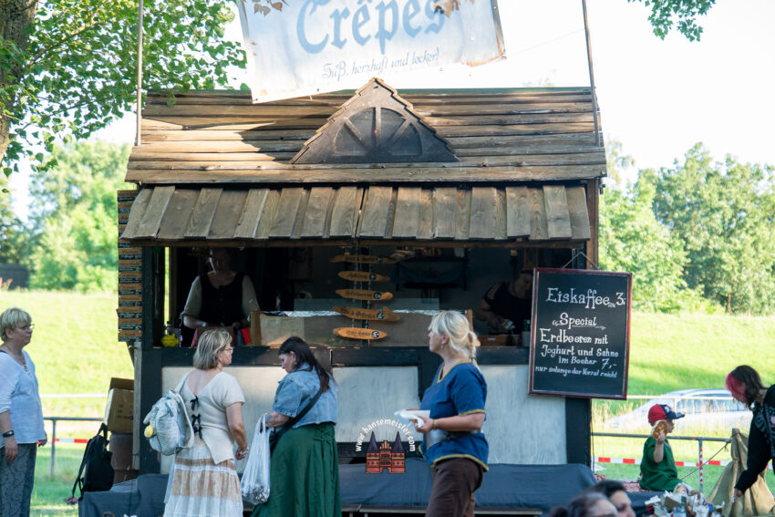 Holzstand für Crêpes auf dem Elb-Spectaculum zu Stove mit Schildern für Speisen und Getränke sowie wartenden Marktbesuchern vor grünem Deichhintergrund.