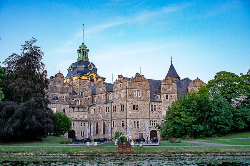 Das Bild MPS-Bückeburg-2-Sa-hansemeister.com (3423).jpg zeigt das majestätische Weserrenaissance-Schloss Bückeburg in seiner vollen Pracht. Die Aufnahme ist vom gegenüberliegenden Ufer der Schlossgräfte aufgenommen, deren ruhiges Wasser den unteren Bildrand prägt. Im Vordergrund erstreckt sich ein schmaler, gepflegter Grünstreifen entlang der Gräfte, auf dem Außengastronomie-Sitzbereiche mit Tischen, Schirmen und Liegestühlen unterhalb der imposanten Fassade platziert sind.

Die historische Schlossfassade aus hellgräulichem Naturstein wird von reich verzierten Giebeln, zahllosen Fenstern und dem markanten grünen Zwiebelturm dominiert, der stolz in den leicht bewölkten, blauen Himmel ragt. Flankiert wird das Bauwerk von dichtem, grünem Baumbestand – links ein prächtiger, dunkler Laubbaum und rechts saftig grüne Schlossgarten-Bäume. Das sanfte Licht fängt die erhabene und friedliche Atmosphäre der Schlosskulisse am Rande des Festivalgeländes perfekt ein.