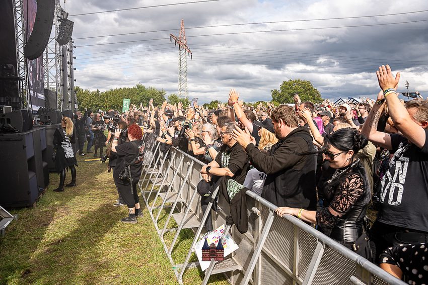 Eine Menschenmenge steht dicht gedrängt in den vorderen Reihen vor einer großen Open-Air-Festivalbühne und applaudiert. Die Zuschauer, viele davon dunkel gekleidet, stehen hinter einer metallischen Sicherheitsbarriere auf einer Rasenfläche. Einige von ihnen haben die Hände zum Klatschen oder Jubeln in die Luft gehoben. Im Hintergrund erstrecken sich grüne Bäume, ein Strommast und ein bewölkter Himmel.