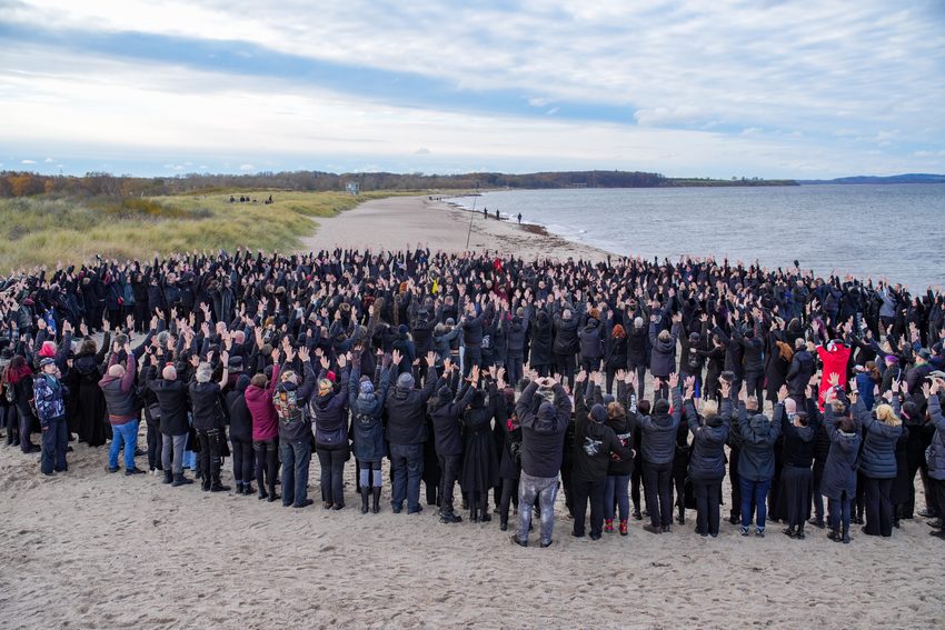 Dieses Foto wurde auf dem Plage Noire Festival 2025 am Weißenhäuser Strand an der Ostsee aufgenommen
Es zeigt eine lange Tradition dieses Musikfestivals der Schwarzen Szene: das alljährliche Gruppenfoto der Besucher am Ostseestrand. Da das Festival traditionell im Frühjahr stattfindet, tragen die überwiegend dunkel gekleideten Festivalbesucher wetterfeste Jacken und Mäntel, während sie gemeinsam für das Foto posieren und die Arme heben.