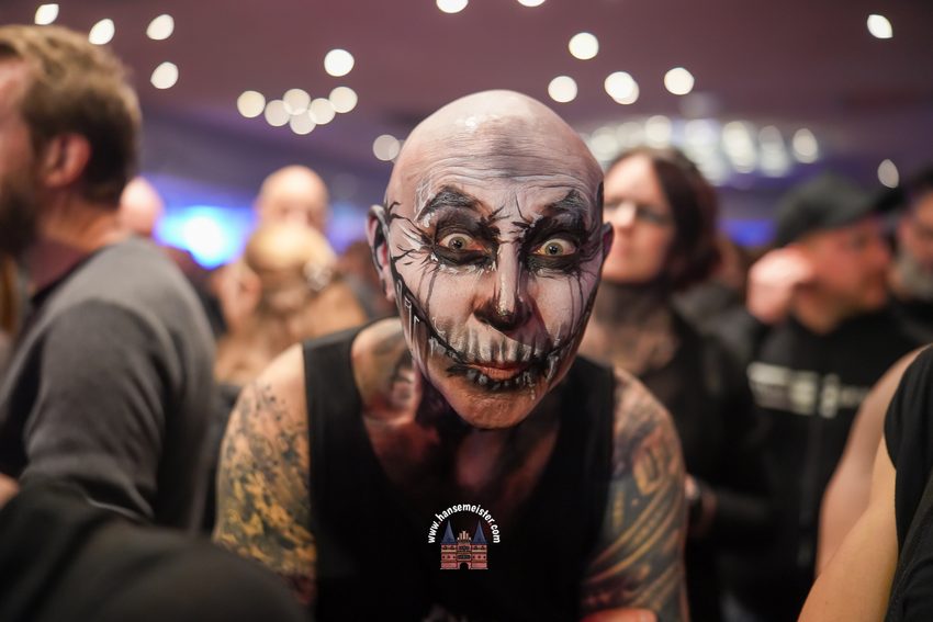 Dieses Foto wurde auf dem Plage Noire Festival 2025 am Weißenhäuser Strand aufgenommen.
Es zeigt einen Festivalbesucher in einer für dieses Gothic-Event typischen, aufwendigen Aufmachung: Ein kunstvolles Skelett- und Totenkopf-Facepainting, das den gesamten Kopf und das Gesicht bedeckt, kombiniert mit tätowierten Armen und einem schwarzen Tanktop.
Das Foto fängt die düstere Eleganz und die detailreichen Stylings ein, die charakteristisch für die Besucher dieses Festivals der Schwarzen Szene sind. Wie bei den vorherigen Bildern stammt die Aufnahme vom Konzert- und Eventfotografen Hansemeister