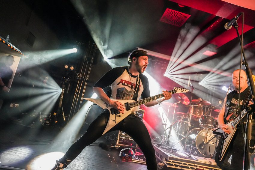 Das Foto zeigt eine energiegeladene Live-Performance zweier Musiker auf einer Konzertbühne, eingefangen in einer dynamischen Weitwinkel-Perspektive.
• Zentraler Musiker (Gitarrist/Sänger): Im Vordergrund steht ein bärtiger Musiker in einer breitbeinigen, rocktypischen Pose. Er trägt eine schwarze Mütze (oder ein Stirnband), ein Baseball-Shirt mit dunklen Ärmeln und einem markanten „Accept“-Band-Aufdruck auf der Brust sowie eine enge schwarze Hose. Mit weit geöffnetem Mund singt oder schreit er voller Emotion, während er eine weiße, pfeilförmige E-Gitarre (V-Form, LTD) spielt.
• Rechter Musiker (Gitarrist): Rechts im Bild ist ein weiterer Gitarrist im Profil zu sehen. Er hat eine Glatze, einen Ziegenbart und trägt eine schwarze Kutte beziehungsweise Weste über einem schwarzen T-Shirt. Auch er spielt eine weiße E-Gitarre mit einer ähnlichen, zackigen Korpusform.
• Hintergrund & Lichtshow: Die Bühne ist in eine spektakuläre Lichtshow getaucht. Mehrere Scheinwerfer strahlen von hinten und den Seiten in den verrauchten Bühnenraum und erzeugen markante, sternförmige Lichtstrahlen (Lens Flares). Auf der rechten Seite sind im Hintergrund Teile des Schlagzeugs (Cymbals) zu erkennen.
• Wasserzeichen: Unten in der Mitte des Bildrandes befindet sich das kreisrunde, rote Logo mit der Aufschrift „hansemeister.com“ als Fotografen-Wasserzeichen.