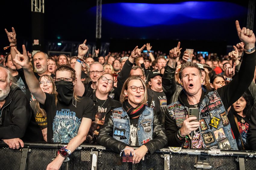 Das Foto zeigt eine begeisterte Zuschauermenge in den ersten Reihen direkt an der Absperrung (Barrikade) eines Metal-Konzerts.
• Vordergrund & Festivalbesucher: Im Fokus stehen mehrere Fans, die dicht aneinandergedrängt mitfeiern. Ein Mann rechts im Vordergrund schaut mit weit geöffnetem Mund und Ohrstöpseln im Ohr begeistert zur Bühne, während er ein Smartphone in den Händen hält. Er trägt eine Jeansweste (Kutte), die über und über mit bunten Heavy-Metal-Aufnähern (unter anderem Iron Maiden und Motörhead) besetzt ist. Neben ihm steht eine lächelnde Frau mit Brille und Schal, die ebenfalls eine mit Patches verzierte Jeansweste über einer Lederjacke trägt. Links von ihr reißt ein Fan mit schwarzem T-Shirt (Metal Hammer-Aufdruck) und schwarzem Tuch vor dem Gesicht den Arm nach oben.
• Publikum & Gesten: Die Menschen im Hintergrund recken enthusiastisch die Arme in die Höhe. Viele von ihnen formen mit den Händen die „Pommesgabel“ (Mano cornuta) oder zeigen mit dem Zeigefinger nach oben. Die Stimmung im dicht gedrängten Publikum ist ausgelassen und fokussiert auf das Geschehen auf der Bühne.
• Hintergrund & Umgebung: Die Szene ist hell ausgeleuchtet, während der obere Hallen- oder Zeltbereich in dunklem Blau gehalten ist. Man erkennt im Hintergrund die tragenden Metallelemente des Zeltes
