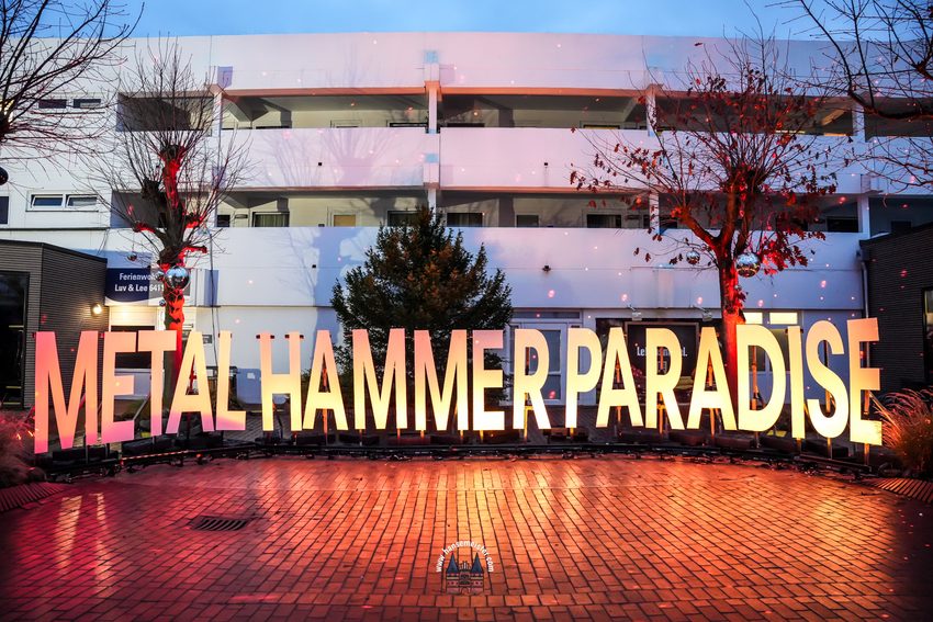 Das Foto zeigt den hell erleuchteten Eingangsbereich des Metal Hammer Paradise Festivals am Weissenhäuser Strand bei Dämmerung.
• Vordergrund & Schriftzug: Im Zentrum steht der riesige, dreidimensionale Schriftzug „METAL HAMMER PARADISE“. Die großen, blockartigen Buchstaben sind von innen heraus in einem warmen Gelb-Orange-Farbton beleuchtet. Sie stehen auf einer Reihe von Scheinwerfern und Halterungen. Der gepflasterte Boden davor glänzt und reflektiert das warme Licht der Buchstaben.
• Hintergrund & Architektur: Direkt hinter dem Schriftzug befindet sich ein mehrstöckiges, weißes Ferienwohnungsgebäude der Ferienanlage (links ist ein Schild mit der Aufschrift „Ferienwohnung Luv & Lee 5411“ zu erkennen). Vor dem Gebäude steht ein grüner Nadelbaum, flankiert von zwei laublosen Bäumen zu den Seiten.
• Beleuchtung & Effekte: Die beiden seitlichen Bäume werden von unten mit intensivem rotem Licht angestrahlt, und in ihren Ästen hängen Discokugeln. Über die gesamte Szene und die weiße Hausfassade sind unzählige kleine, rote Lichtpunkte eines Lasereffekts gestreut.